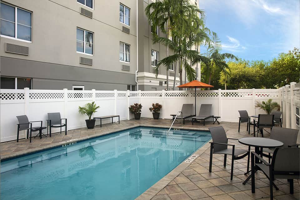 Hotel courtyard pool with loungers, white privacy fence, palm trees, and modern building facade