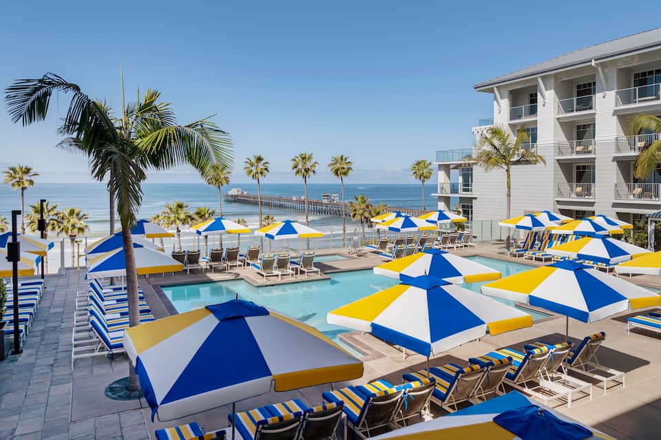 Beachfront resort pool area with blue and yellow umbrellas, loungers, palm trees, ocean view, and white building