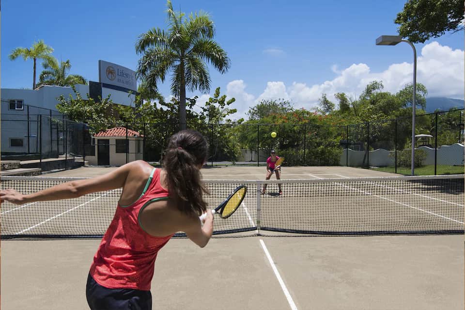 Woman playing tennis on outdoor court with mountains visible in background and palm trees