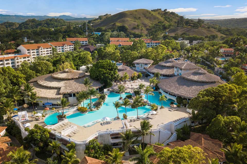 Aerial view of tropical resort with turquoise pool, thatched-roof structures, palm trees, and surrounding mountains