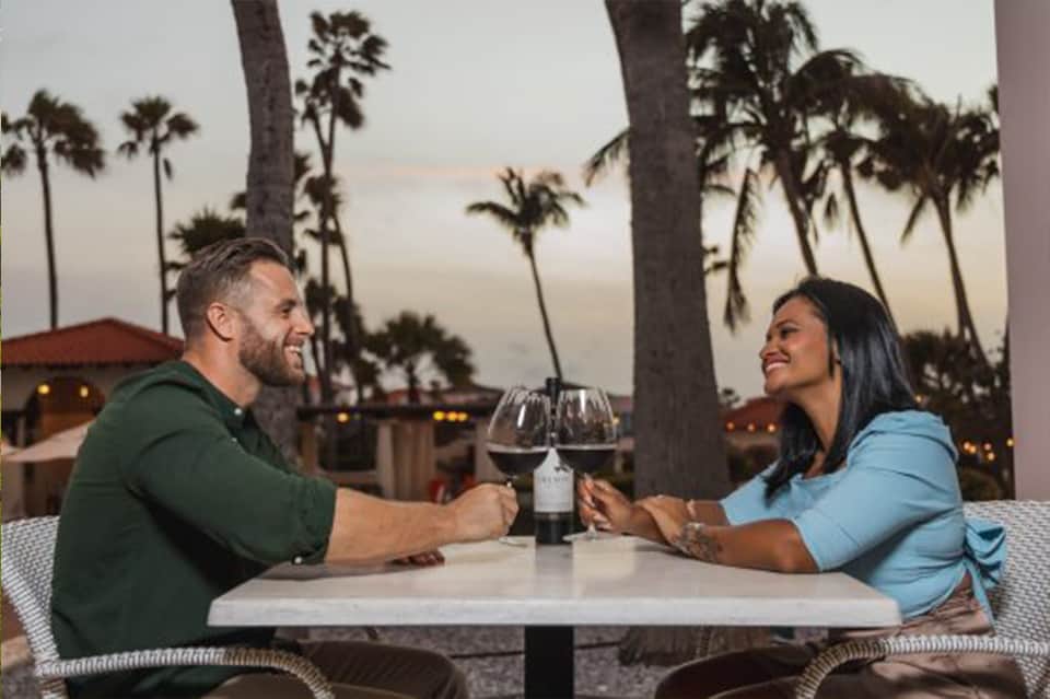 Couple enjoying wine at outdoor dining table at sunset with palm trees in background