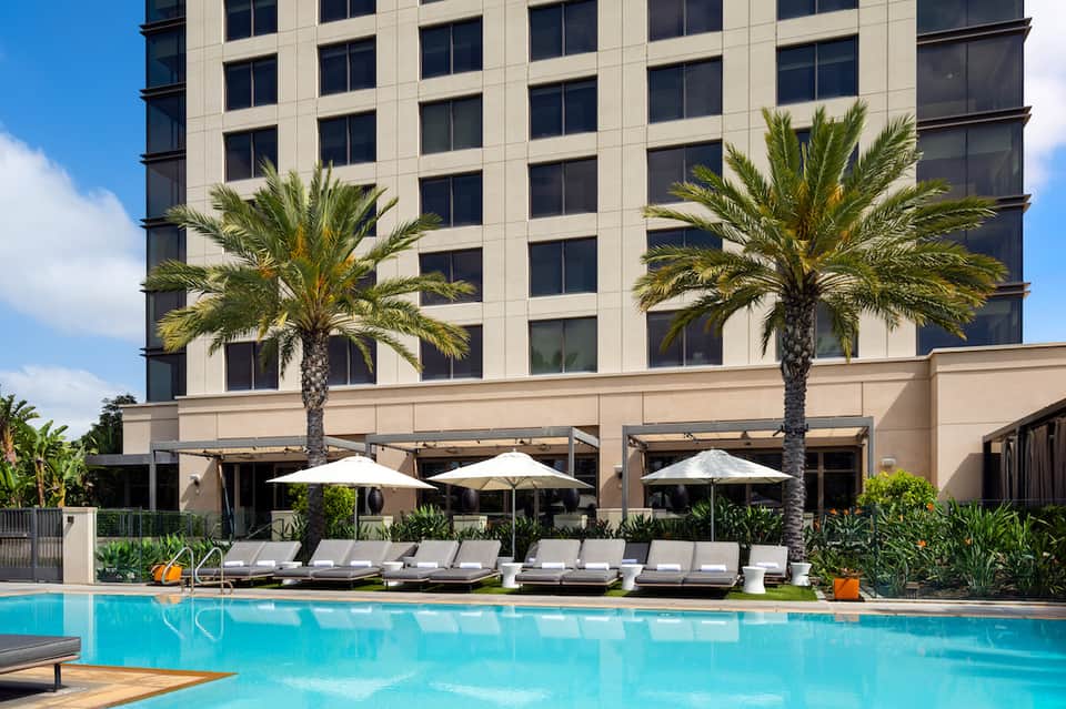 Resort pool deck with lounge chairs, umbrellas, palm trees, and modern hotel tower building