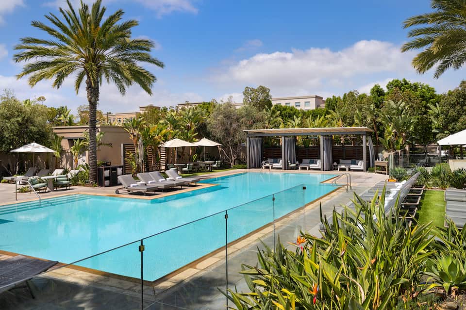 Resort pool surrounded by palm trees, loungers, umbrellas, and modern pavilions