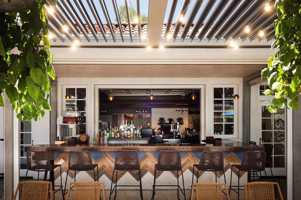 Modern outdoor bar area with wooden pergola ceiling, pendant lights, bar seating, and shelving displaying bottles and glassware