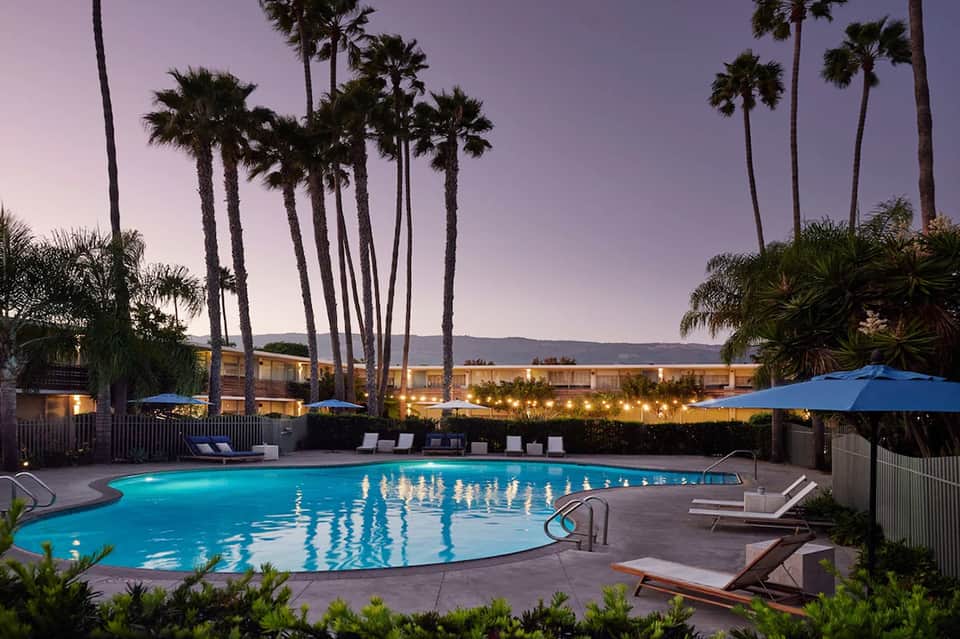 Lit swimming pool at dusk surrounded by palm trees and blue umbrellas with resort buildings in background