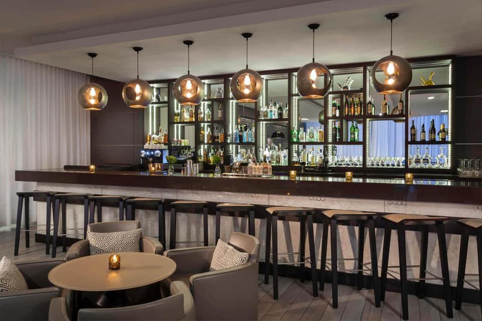 Hotel bar with copper pendant lights, black bar seating, and shelves of liquor bottles