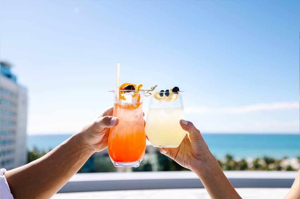 Two tropical cocktails toasting against ocean and sky backdrop with decorative fruit garnishes