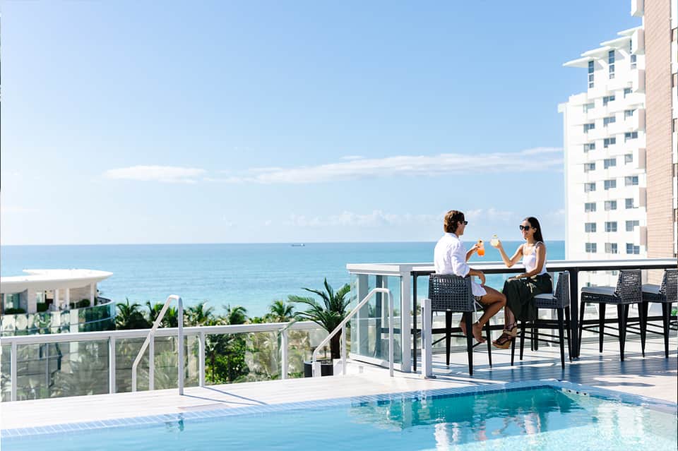 Beachfront hotel pool deck with ocean view, two guests dining at table, high-rise building, tropical plants