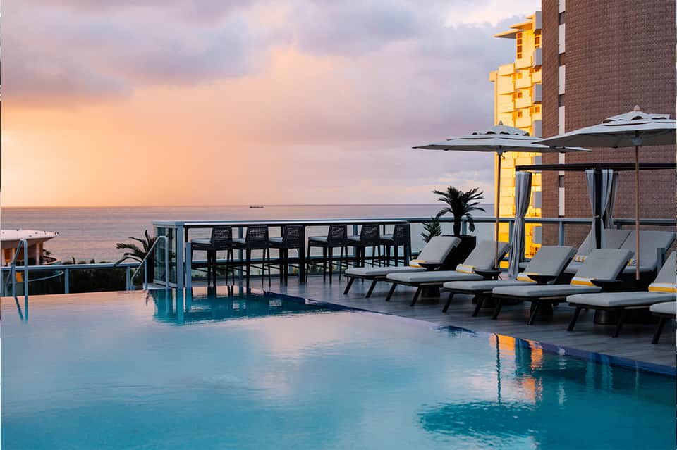 Rooftop pool at sunset with lounge chairs, umbrellas, and ocean view backdrop