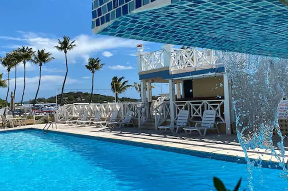 Resort pool with turquoise water, white railings, palm trees, and tropical beach bar with ocean views
