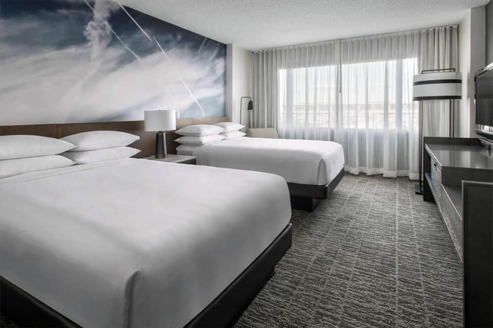 Modern hotel room with two queen beds, floor-to-ceiling windows, and cloud artwork on accent wall