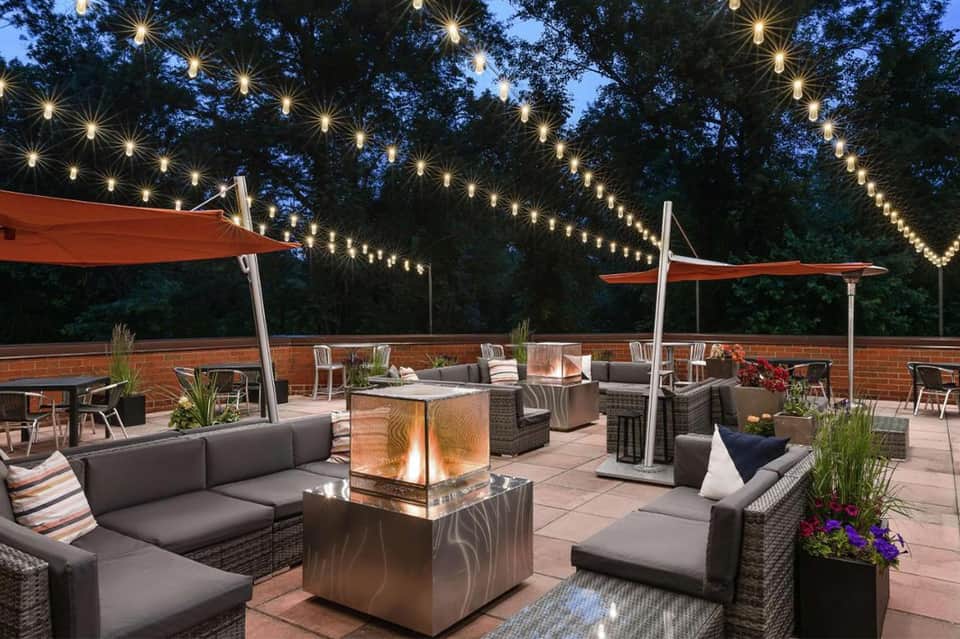 Nighttime rooftop patio with string lights, fire pit, lounge seating, and red umbrellas