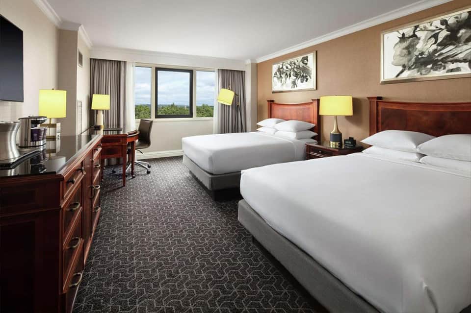 Upscale hotel room with two queen beds, wooden furniture, and panoramic window view of trees