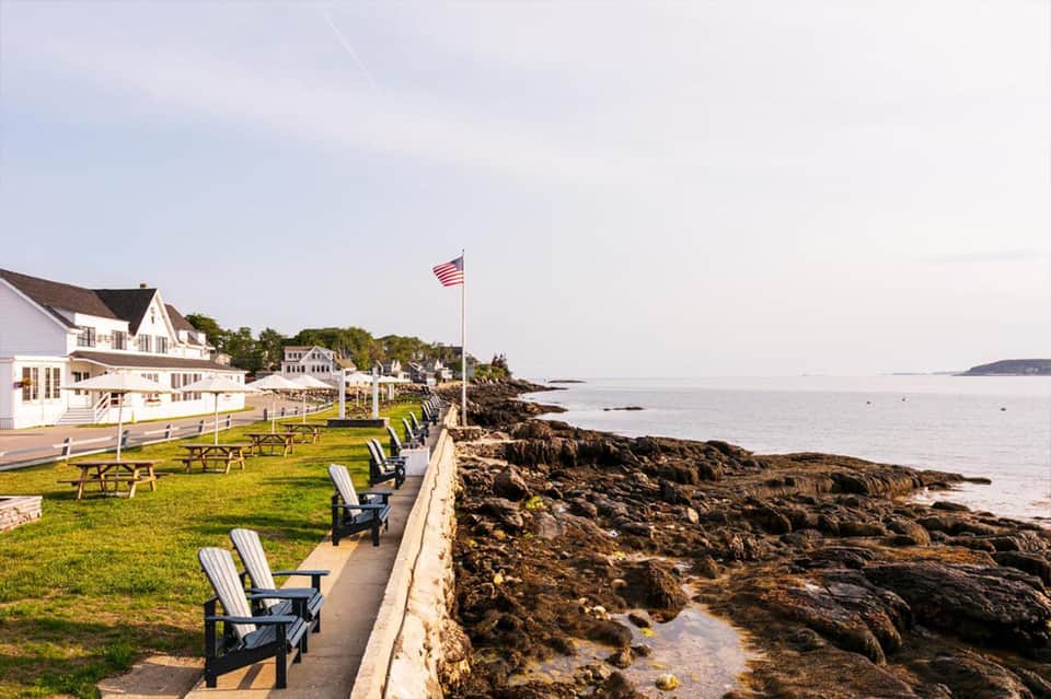 Coastal waterfront with Adirondack chairs on lawn, white buildings, rocky shoreline, and ocean view