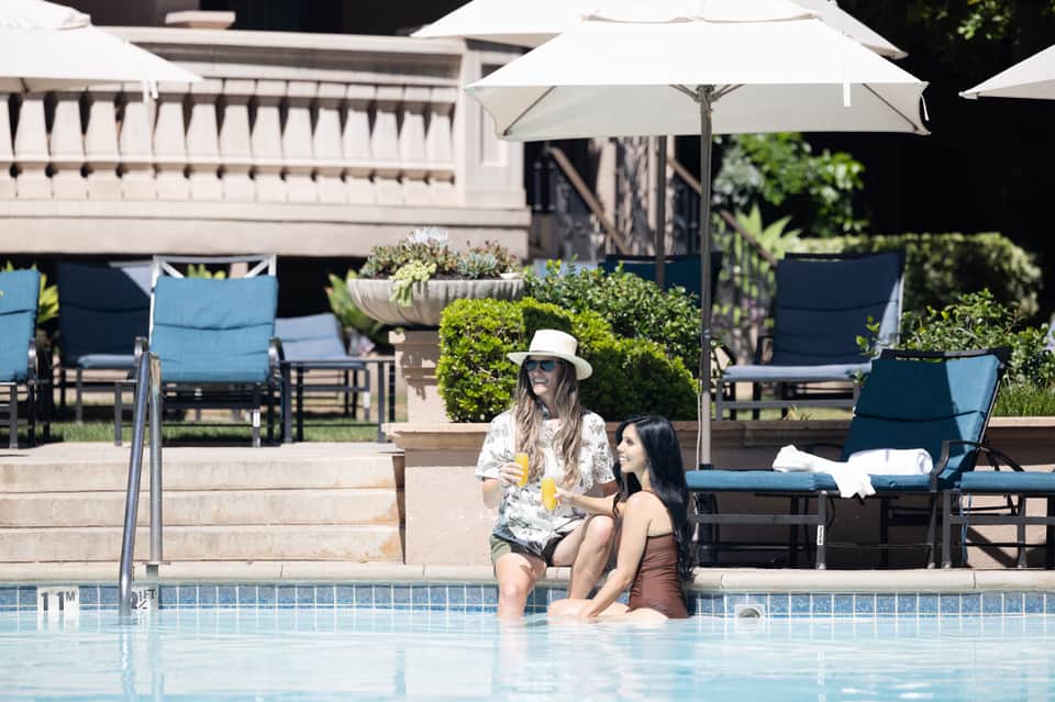 Two women relaxing in shallow pool water with white umbrellas and blue loungers in background