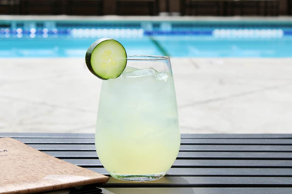 Cucumber-garnished cocktail on poolside table with swimming pool in background