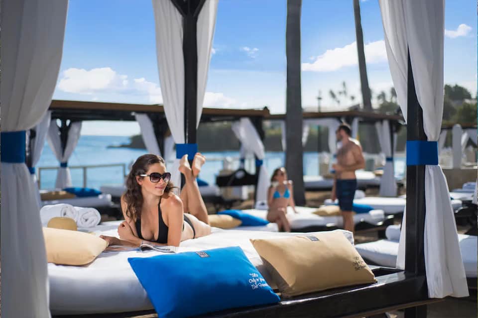 Woman relaxing on a beachside cabana bed with white curtains and water views