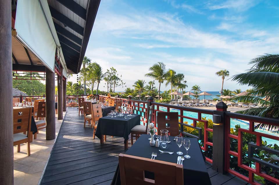 Waterfront dining terrace with ocean views, palm trees, and coastal resort architecture