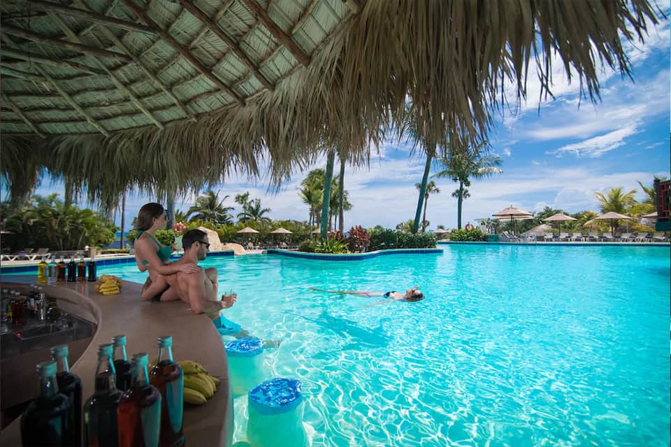 Resort guests relaxing by tropical lagoon pool with thatched roof shelter and palm trees