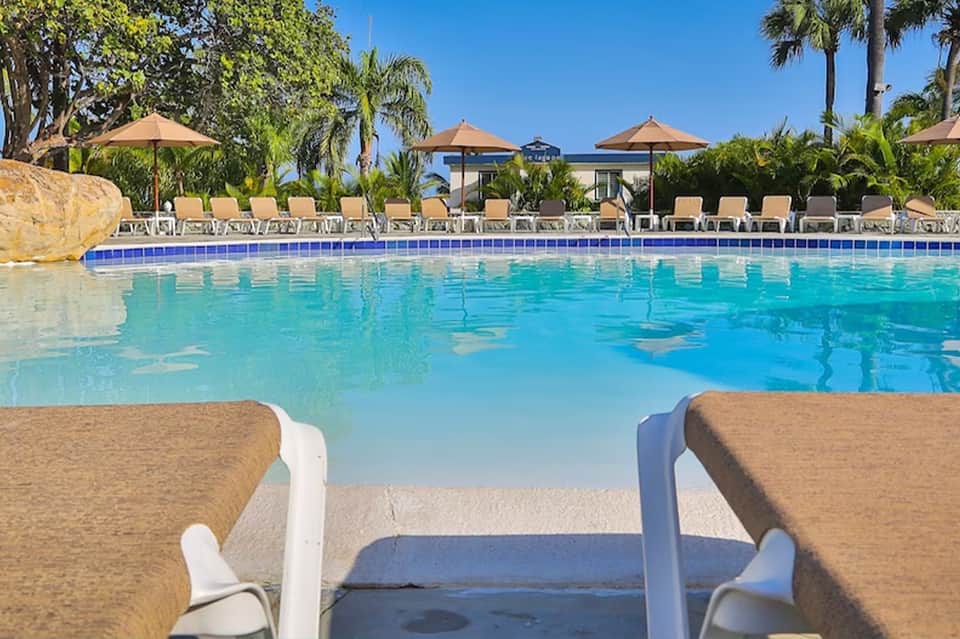 Resort swimming pool with blue water, lounge chairs, umbrellas, and palm trees under clear sky