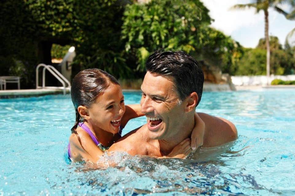 Father and daughter swimming together in bright blue resort pool with tropical landscaping