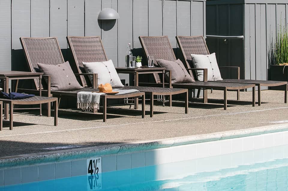 Poolside lounge area with woven chairs, cushions, and small tables beside a bright blue swimming pool