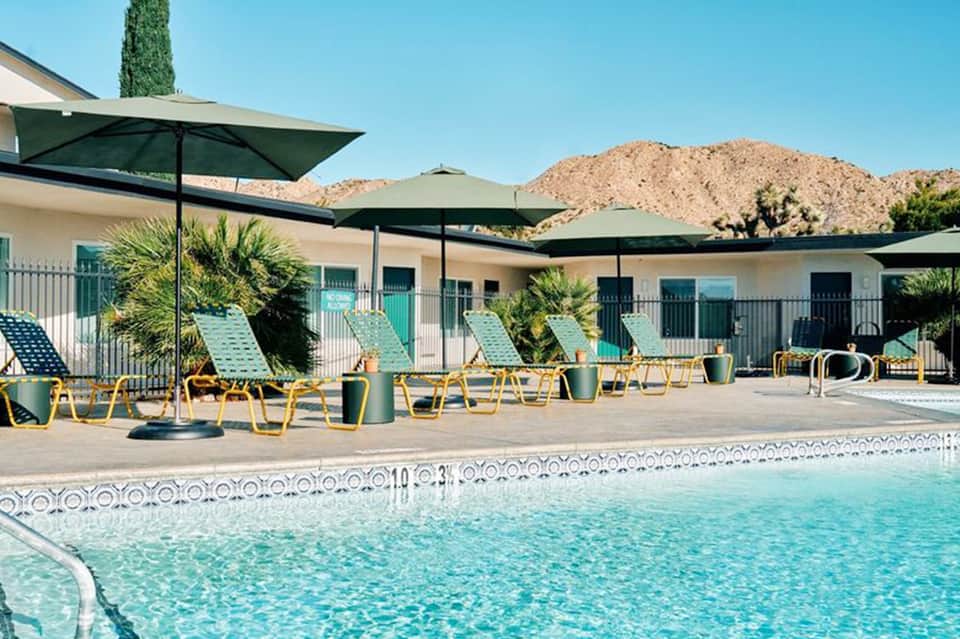Resort pool area with turquoise water, green umbrellas, yellow loungers, palm trees, and desert mountains in background