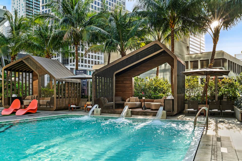 Urban rooftop pool with wooden cabanas, palm trees, and turquoise water surrounded by high-rise buildings