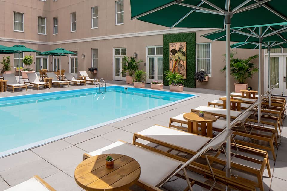 Resort courtyard with blue swimming pool, wooden loungers, green umbrellas, and beige building
