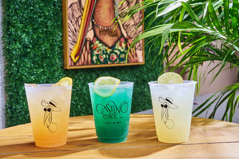 Three colorful cocktails with lime garnish displayed on wooden counter with green wall backdrop
