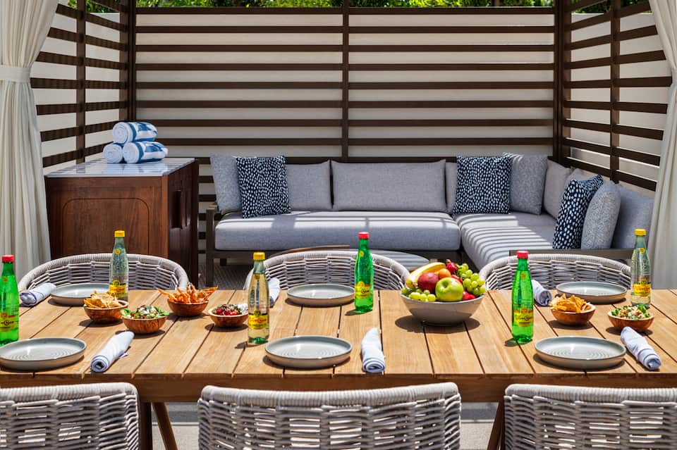 Upscale outdoor dining setup with striped privacy screen, gray seating, wooden table, and fresh fruit display
