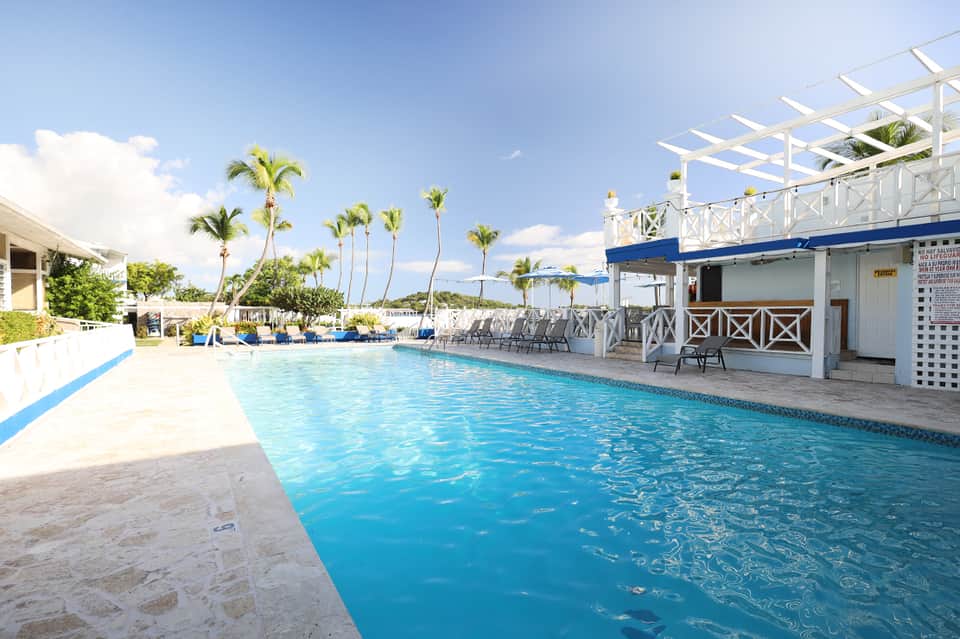 Tropical resort outdoor swimming pool with palm trees, white lounge chairs, and blue cabana structures under clear sky