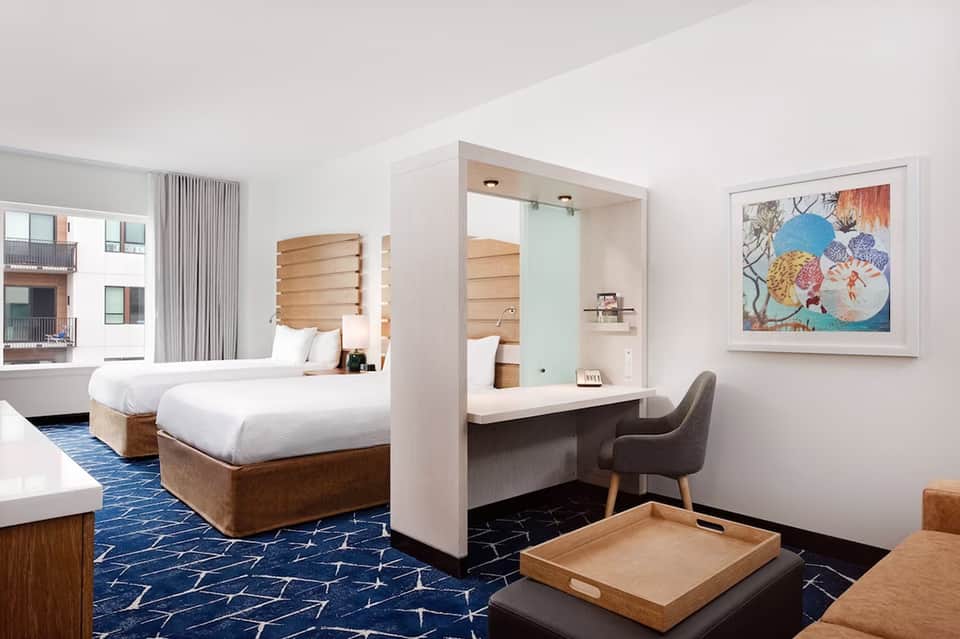 Modern hotel room with two queen beds, wooden headboards, desk with chair, artwork on wall, blue patterned carpet