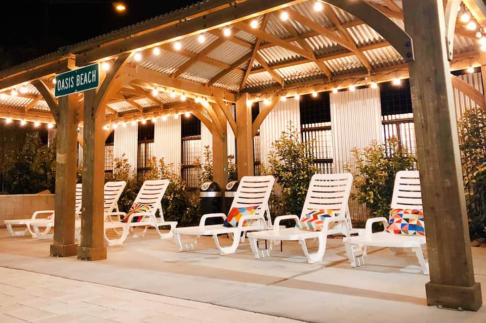 Wooden pergola with string lights, white lounge chairs, and Oasis Beach signage