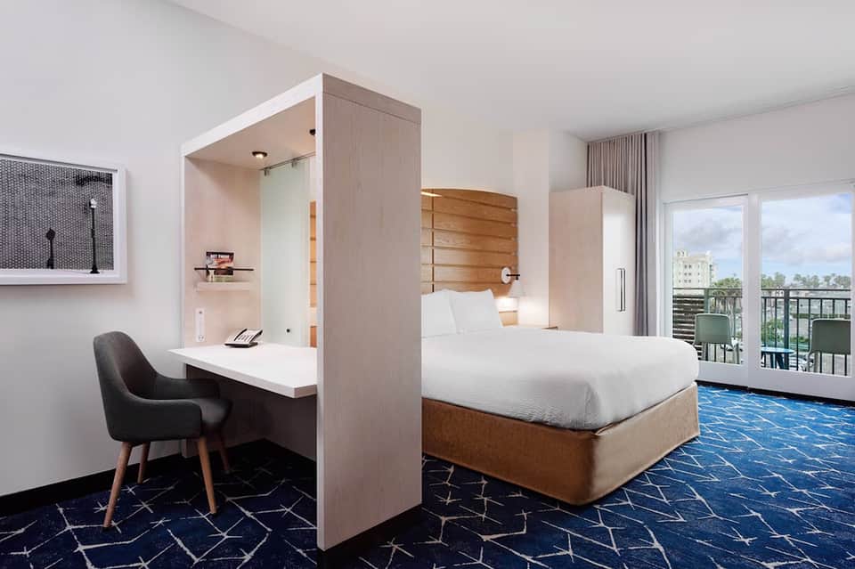 Contemporary hotel bedroom with wooden headboard, desk nook, and balcony with city views