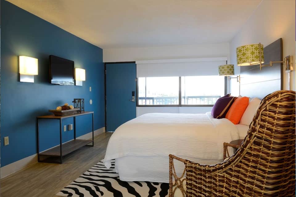 Hotel bedroom with blue accent wall, white bedding, colorful pillows, and balcony view