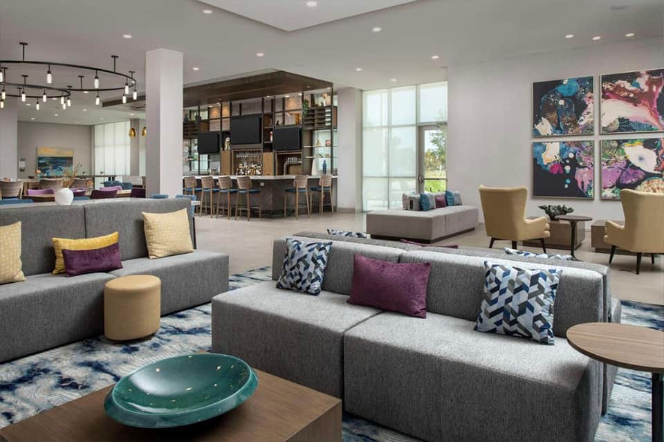 Modern hotel lobby with gray sectional sofas, colorful artwork, bar area, and large windows overlooking grounds