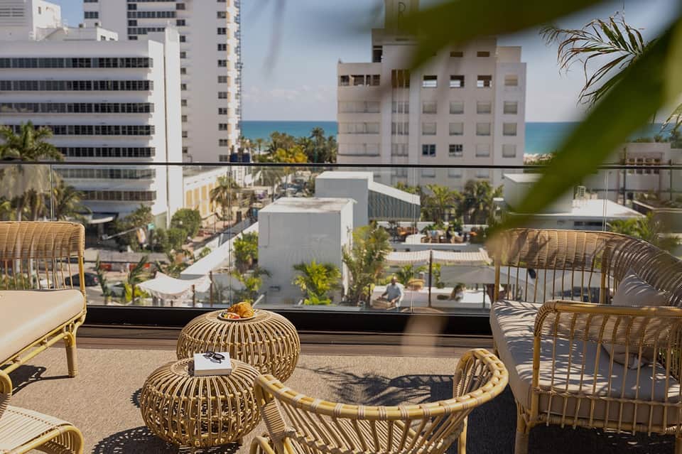 Beachfront balcony with wicker seating overlooking ocean, palm trees, and coastal buildings