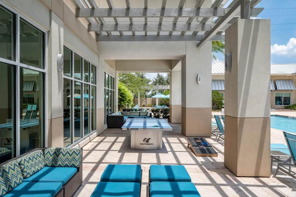 Modern covered patio with cornhole game, lounge seating, and view of pool area beyond