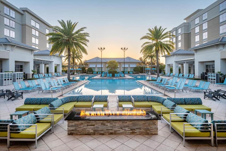 Resort pool at sunset with turquoise water, lounge chairs, firepit, and palm trees flanking modern buildings