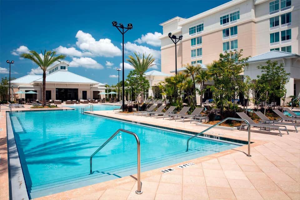 Resort pool area with palm trees, lounge chairs, and pink hotel building under blue sky