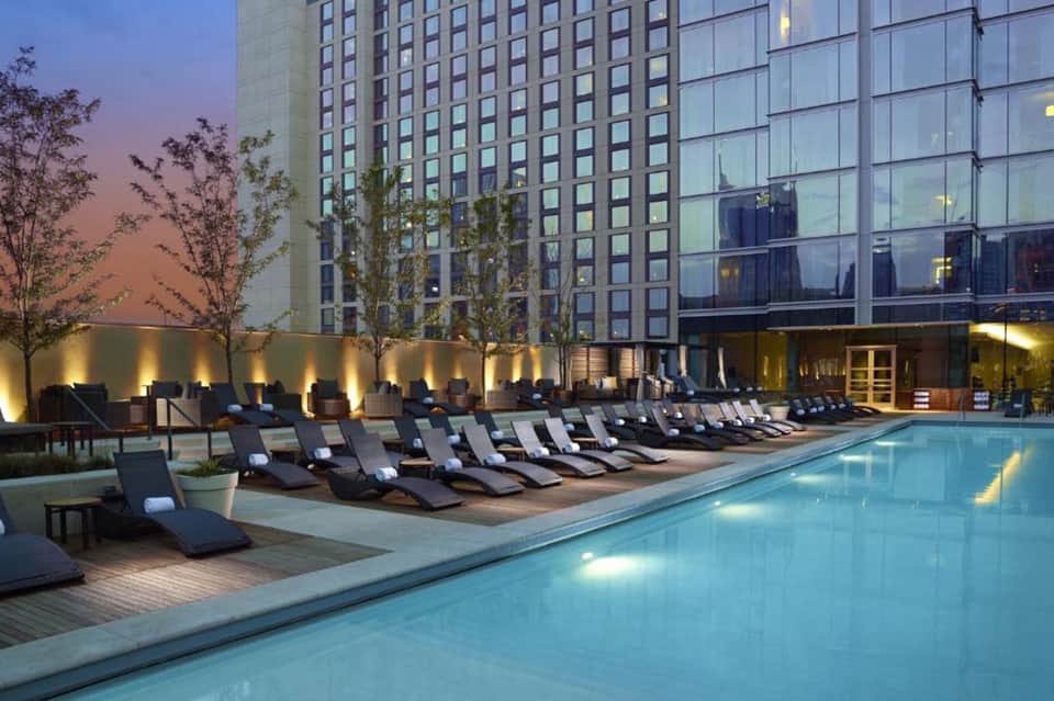 Rooftop pool at dusk with lounge chairs, illuminated high-rise hotel building, and city views