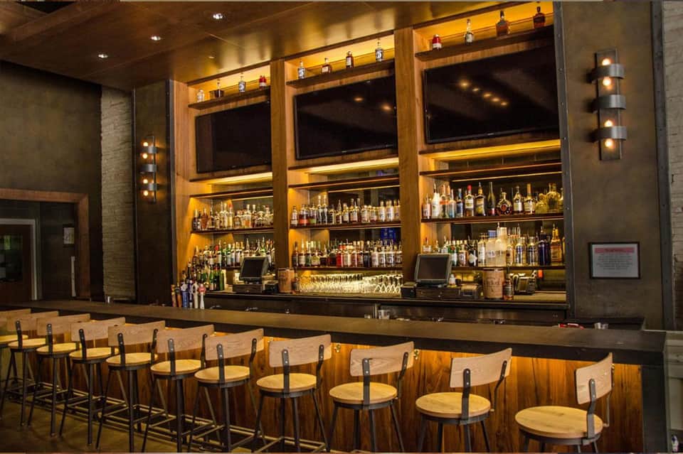 Upscale bar with wooden counter, illuminated shelving displaying spirits, and comfortable seating