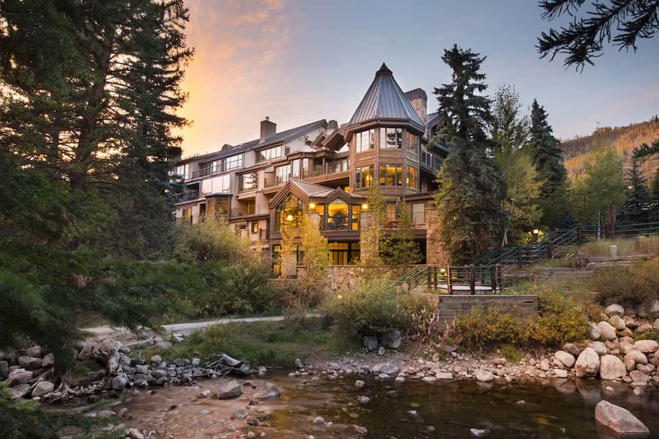 Luxury mountain lodge by stream with stone architecture, turret, and autumn foliage at golden hour