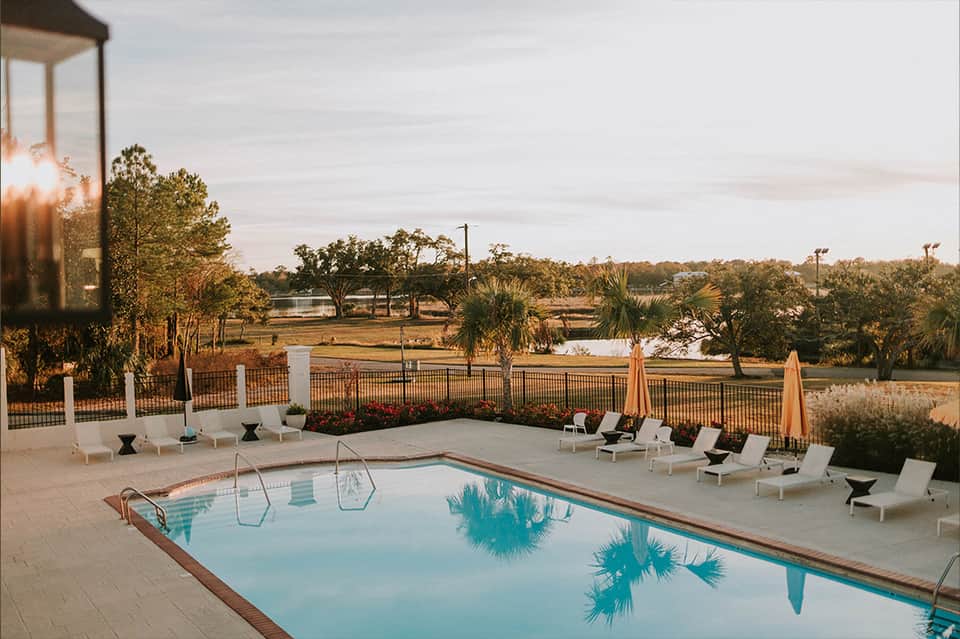 Lakeside pool at dusk with white lounge chairs, umbrellas, fence railing, and trees reflected in calm water