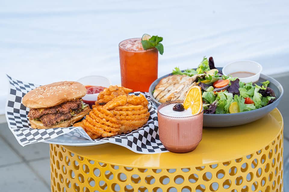 Gourmet burger, waffle fries, grilled chicken salad, and cocktails served on yellow table with ocean view