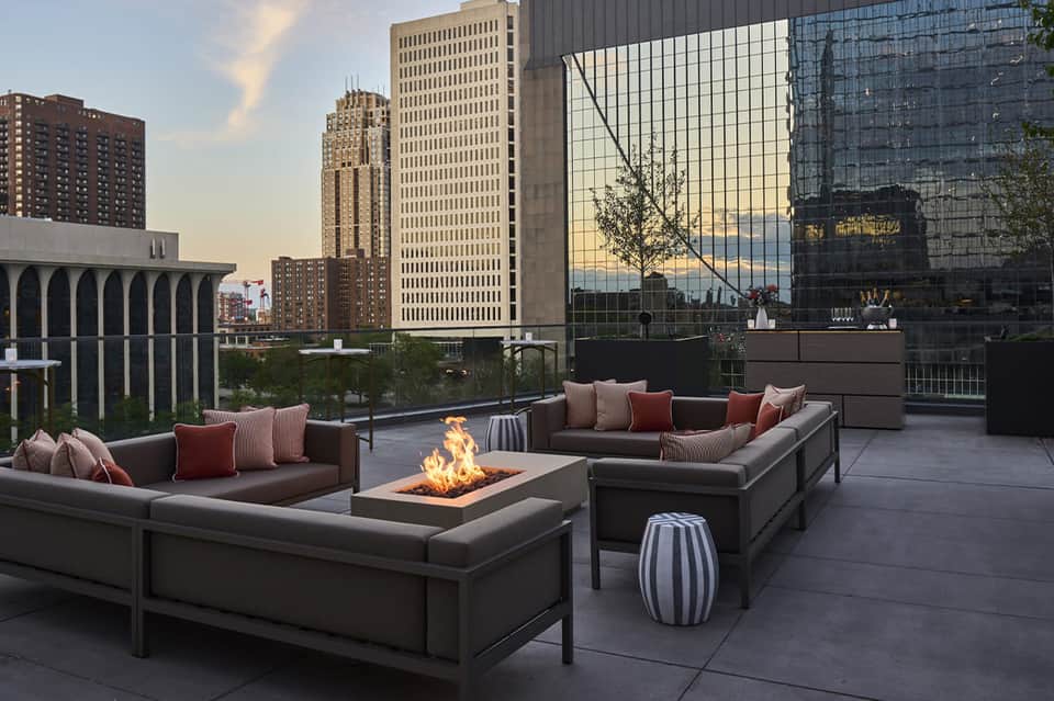 Rooftop lounge with fire pit, gray seating, city skyline views, and downtown buildings at dusk