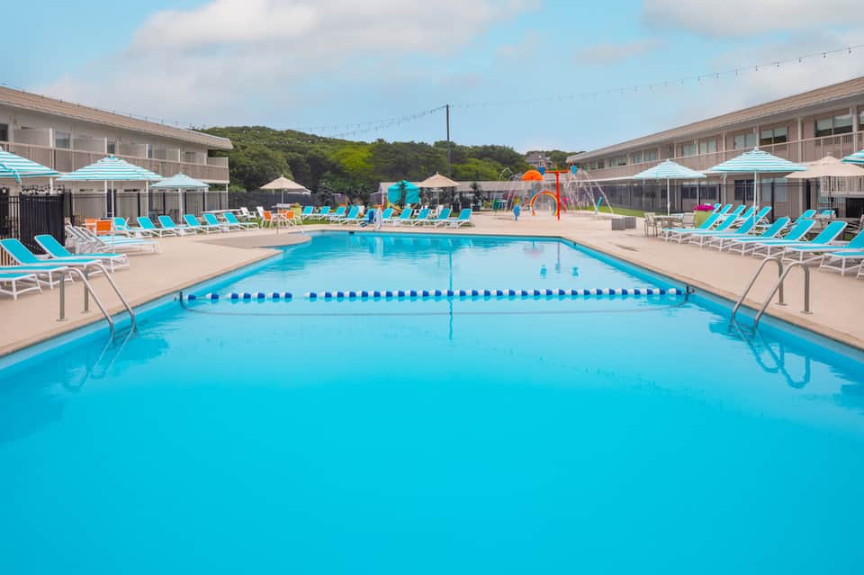 Bright outdoor swimming pool with turquoise water, lane dividers, lounge chairs, and playground