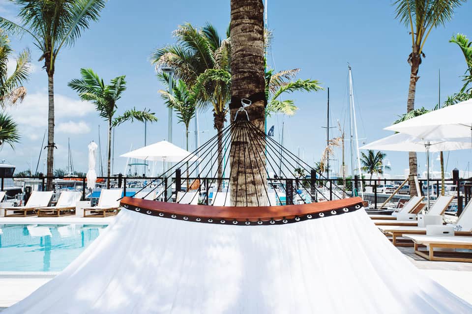 Luxury poolside hammock suspended from palm tree with white umbrellas and sailboats in background