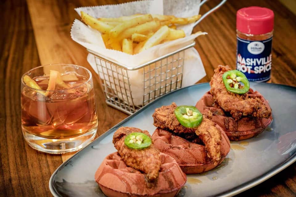 Fried chicken sandwich with jalapeños served with fries, bourbon drink, and hot sauce on wooden table